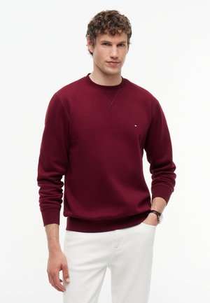 Hombre con cabello rizado que lleva una sudadera color vino y pantalones blancos, de pie con una mano en el bolsillo contra un fondo blanco.