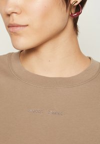 Beige Baumwoll-T-Shirt mit Rundhalsausschnitt, das den gestickten Text "SAMSMØE" zeigt. Nahaufnahme, die die glatte Textur und die subtilen Nähdetails hervorhebt.
