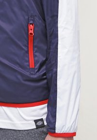 Námořnická modrá a bílá bunda se zipovou kapsou, červeným zipem a lemováním, hladkou texturou a elastickým pasem a manžetami.