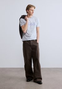 Ανοιχτό μπλε μπλουζάκι με τη λέξη "BOSS" σε μοβ χρώμα, συνδυασμένο με καφέ παντελόνι φαρδιάς γραμμής και σκούρο πουλόβερ κρεμασμένο σε έναν ώμο. Μαύρα παπούτσια.