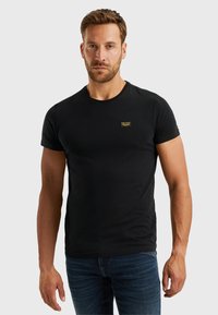 Ein erwachsener Mann mit kurzen braunen Haaren, der ein eng anliegendes schwarzes Kurzarm-T-Shirt und blaue Jeans trägt, steht vor einem schlichten hellen Hintergrund.
