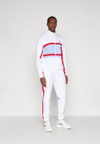 Λευκή φόρμα με κόκκινες και ανοιχτό γαλάζιες οριζόντιες ρίγες. Διαθέτει τζάκετ με φερμουάρ και παντελόνι με ελαστική μέση. Συνδυάζεται με λευκά αθλητικά παπούτσια.