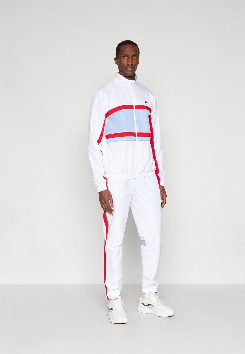 Λευκή φόρμα με κόκκινες και ανοιχτό γαλάζιες οριζόντιες ρίγες. Διαθέτει τζάκετ με φερμουάρ και παντελόνι με ελαστική μέση. Συνδυάζεται με λευκά αθλητικά παπούτσια.