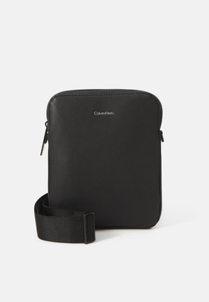 Saco de ombro preto feito de material sintético texturizado. Apresenta um fecho com zíper e uma alça ajustável. Pequeno logo na parte da frente. Forma quadrada.