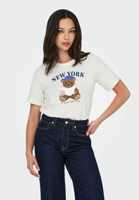 Cremefarvet bomulds T-shirt med et grafisk tryk af en bjørn iført en tanktop og en pink drink, samt teksten "NEW YORK City of Dreams" i blåt.