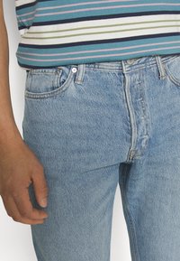 Jeans in denim azzurro chiaro con taglio dritto, dotati di chiusura a bottone, passanti per cintura e design a cinque tasche. Top a righe visibile sopra la vita.