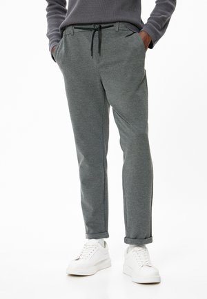 Pantalon de survêtement gris avec un cordon de serrage à la taille, poches latérales et coupe fuselée, associé à des baskets blanches sur un fond uni.