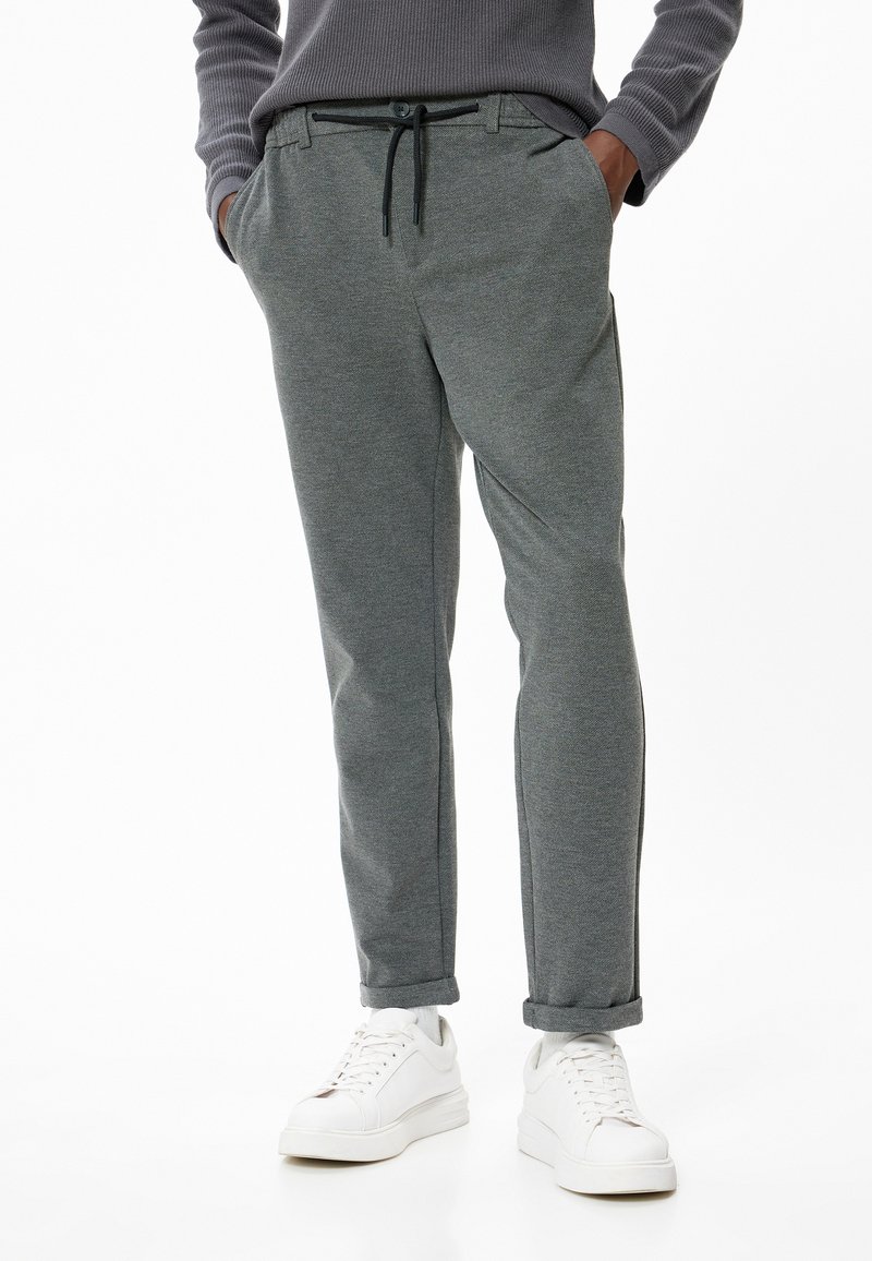 Pantaloni sportivi grigi con vita regolabile tramite cordoncino, tasche laterali e vestibilità affusolata, abbinati a sneakers bianche su uno sfondo semplice.