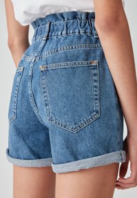 Persona vestida con pantalones cortos de mezclilla azul de cintura alta y dobladillo enrollado, y una blusa blanca, vista desde la parte baja de la espalda hasta la mitad del muslo.