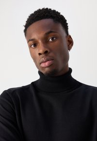 Suéter de cuello alto negro hecho de tela elástica, con un diseño ajustado. El sujeto tiene el cabello corto y texturizado y lleva un pequeño pendiente.