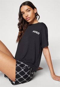 T-shirt noir à manches courtes avec le logo "HUGO" en blanc, associé à un short à motifs présentant un design en maillons de chaîne blanc sur fond noir.