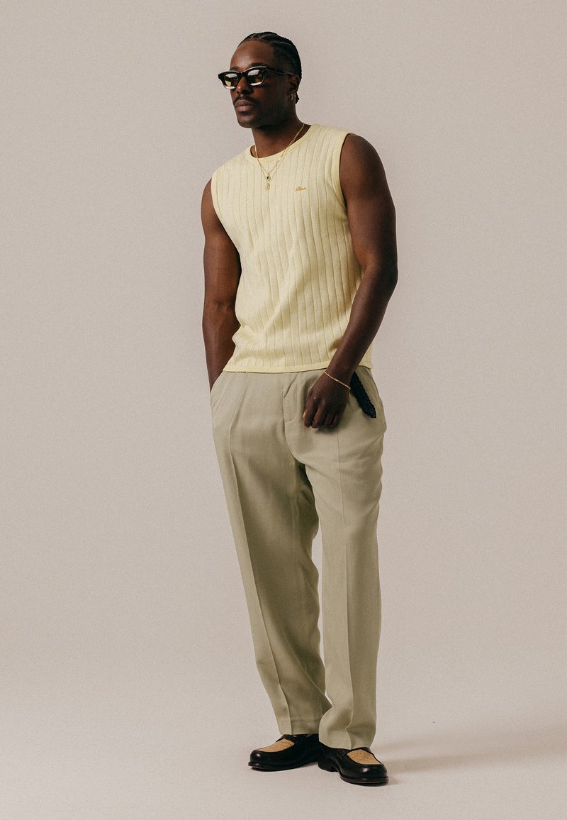 Homme aux cheveux tressés et portant des lunettes, vêtu d'un débardeur jaune côtelé, d'un pantalon beige et de chaussures bicolores noires et beige, debout devant un fond uni.