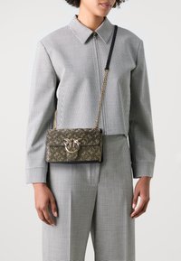 Piccola borsa rettangolare con un motivo ornato in oro e nero, caratterizzata da un accento metallico circolare e una tracolla a catena, indossata a tracolla.