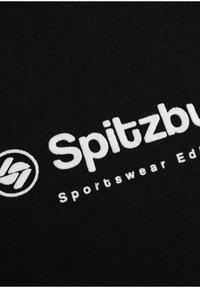 Black fabric featuring white embroidered text: "Spitzbuh Sportswear Edition," with a circular logo to the left of the text.