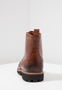 Botín de cuero marrón con detalles de brogue, panel lateral elástico y asa. Suela negra de goma resistente con surcos para tracción.