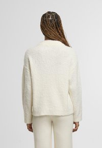 Žena s dlouhými copy, oblečená v bílém huňatém svetru a odpovídajících kalhotách, zobrazená zezadu na jednobarevném pozadí.