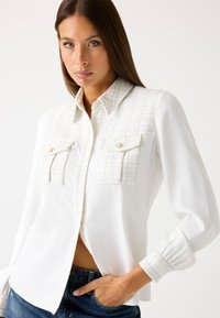 Weißes Bluse mit strukturiertem karierten Muster an der Passe, zwei vorderen Taschen und Knopfverschluss. Mit Puffärmeln und Manschettendetails.