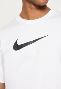Bílý bavlněný tričko s výrazným černým logem Nike swoosh. Tkanina vypadá hladce a má klasický design s kulatým výstřihem.