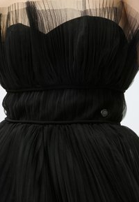 Μαύρο strapless φόρεμα με στρωτό διαφανές τούλι, μαζεμένο στη μέση, και ένα μικρό στρογγυλό μεταλλικό έμβλημα στο ισχίο.