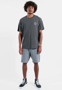 T-shirt en coton gris foncé à manches courtes avec un logo circulaire sur la poitrine, associé à un short gris clair et des baskets montantes noires.