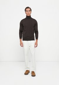 Man standing against a white background wearing a dark brown turtleneck sweater, white pants, and brown suede shoes.