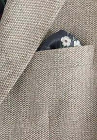 Blazer de tweed beige con un patrón de espiga texturizado; presenta un pañuelo de bolsillo azul marino con un diseño floral asomando del bolsillo del pecho.