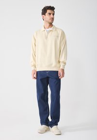 Sudadera de cremallera de color beige claro con cuello alto, combinada con vaqueros de color azul oscuro y zapatillas de color claro. Tejido suave con un corte relajado.