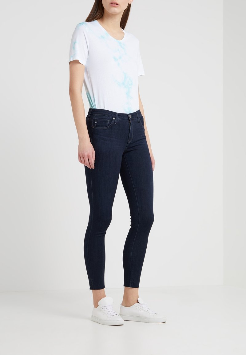 Vit kortärmad t-shirt med blå tie-dye detaljer, tillsammans med mörkblå åtsittande jeans och vita sneakers. Enkelt design, mjuk textur.