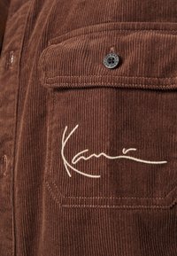 Camicia in tessuto di velluto a coste marrone con taschino frontale, caratterizzata da una firma ricamata bianca e dettagli in bottoni a pressione neri. Texture a coste verticali.