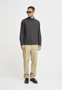 Dark grey button-up shirt with long sleeves, beige chinos, and black shoes. Model wearing dark sunglasses, standing on a plain background.