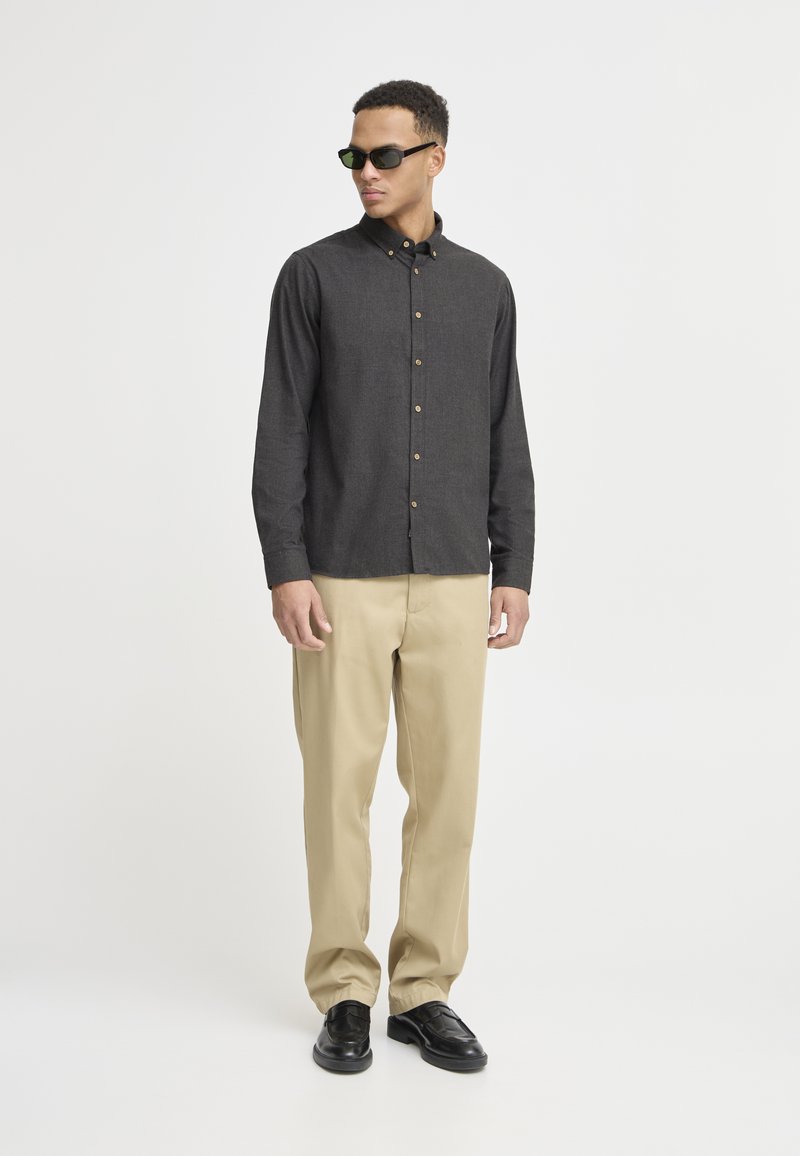 Dunkelgraues Hemd mit langen Ärmeln, beigen Chinos und schwarzen Schuhen. Model trägt eine dunkle Sonnenbrille und steht vor einem einfarbigen Hintergrund.