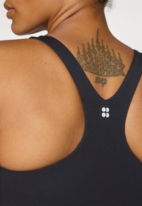 Črn športni modrček z dizajnom racerback, ki ima gladko teksturo in majhen bel logotip na hrbtu. Vidna je tudi koža.