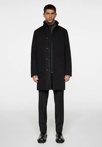 Fekete külső kabát cipzárral és patentgombokkal, szürke pulóver fölött viselve. Egyenes szabású, hosszú ujjú. Sima textúrájú.