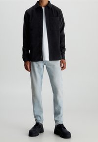 Černá kordová košile na tlačítka s jednou kapsou, nošená přes bílý tričko, zkombinovaná se světlými modrými džínami a černými platformovými botami.