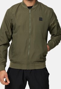 Homme portant une veste bomber vert olive avec fermeture éclair devant, poignets et ourlet côtelés, et pantalon noir, main partiellement dans la poche.