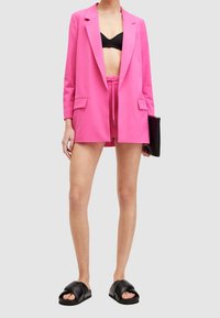 Rosa skräddarsydd kavaj med slag, ihop med en svart crop-top, matchande shorts och svarta sandaler, mot en neutral bakgrund.