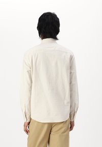 Lys beige skjorte med knapper lavet af tekstureret bomuld. Har lange ærmer, en standard krave og en buet kant. Ingen synlige mønstre.