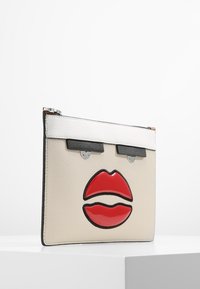 Krémová kožená psaníčko s lesklým designem červených rtů, stříbrným kováním a černými akcenty. Obdélníkový tvar, texturovaný povrch a uzávěr na zip.