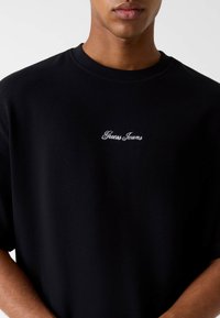 Czarny T-shirt z krótkim rękawem wykonany z prążkowanej tkaniny, z haftowanym białym logo "Guess Jeans" na piersi. Prosty i czysty design.