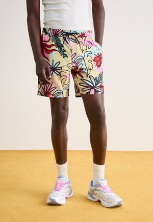 Pantalones cortos multicolores con un diseño floral y abstracto; cuentan con una cinturilla elástica con cordón. Llevados con zapatillas deportivas gris claro.