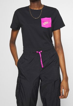 Personne portant un t-shirt Nike noir avec une poche poitrine rose vif et un cordon assorti rose sur un pantalon noir, collier chaîne en or visible.