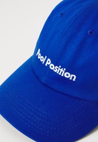 Căciulă de baseball albastră din bumbac, cu vârful structurat. Prezintă un text brodat alb "Pool Position" pe față și o textură subtilă.