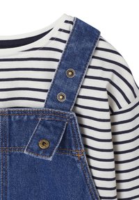 Jeanslatzhosen mit verstellbaren Trägern und goldfarbener Hardware. Darunter befindet sich ein weiß-blau gestreiftes Langarmshirt.