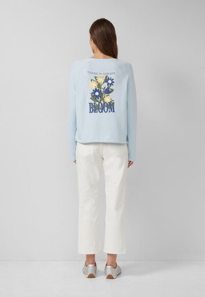 Vrouw die met de rug naar de camera staat, draagt een lichtblauw sweatshirt met bloemenprint en tekst "There is always time to bloom", witte cropped broek en zilveren sneakers.