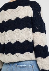 Strickpullover mit abwechselnd navyblauen und cremefarbenen Zickzackstreifen, getragen mit hellblauen Denim-Jeans.