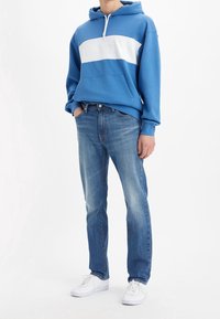 Sudadera azul con franjas y logotipo blancos, con bolsillo delantero, combinada con vaqueros azules deslavados. El modelo lleva zapatillas blancas. Un atuendo casual y sencillo.