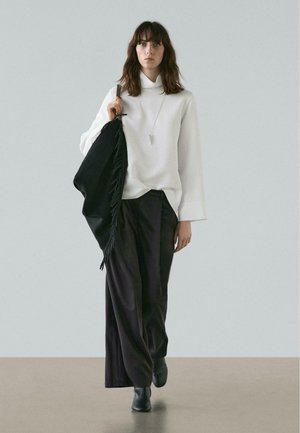 Maglione bianco a collo alto, sciarpa nera con frange, pantaloni larghi scuri e stivaletti neri, in piedi su un pavimento beige davanti a uno sfondo grigio.