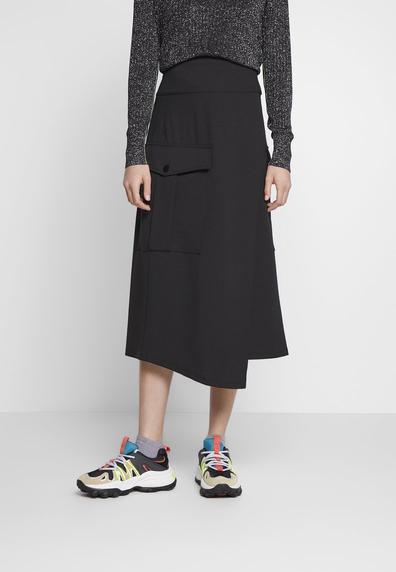 Falda negra de longitud a la rodilla con un dobladillo asimétrico, dos grandes bolsillos delanteros y una cinturilla elástica, combinada con zapatillas deportivas coloridas.