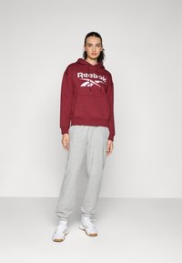 Felpa bordeaux con logo Reebok bianco, tasca frontale, abbinata a pantaloni da tuta grigi chiari e scarpe da ginnastica bianche con suola in gomma.