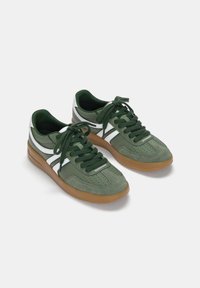 Une paire de baskets vertes avec des rayures blanches, des lacets verts et des semelles en caoutchouc gomme, présentées côte à côte sur un fond blanc.